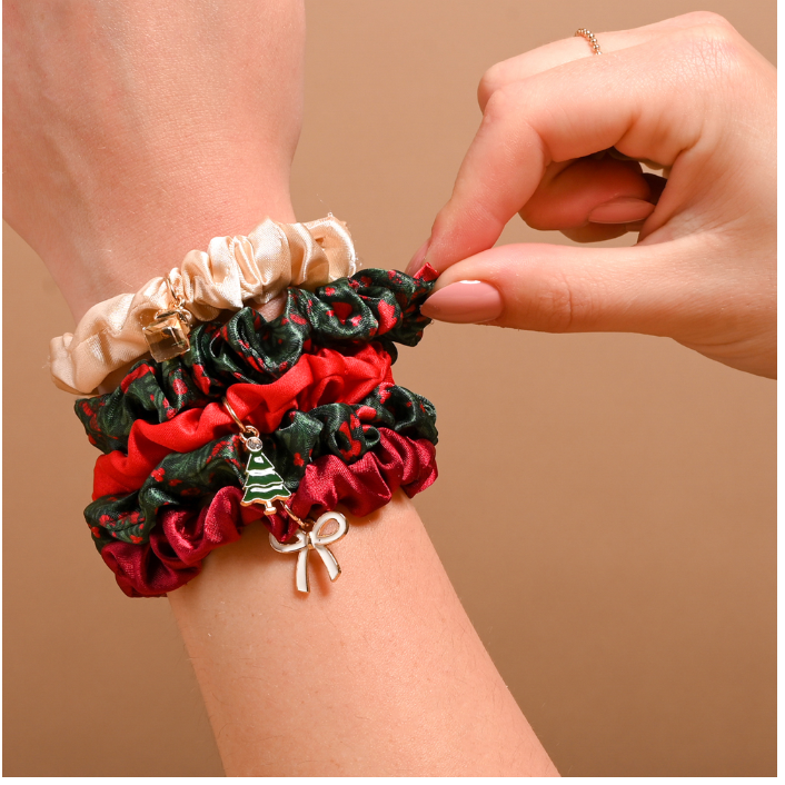 A close-up of a person's wrist wearing stacked fabric scrunchie bracelets in cream, dark green, and red, with small holiday-themed charms including a miniature tree and bow. A hand is adjusting the top bracelet.