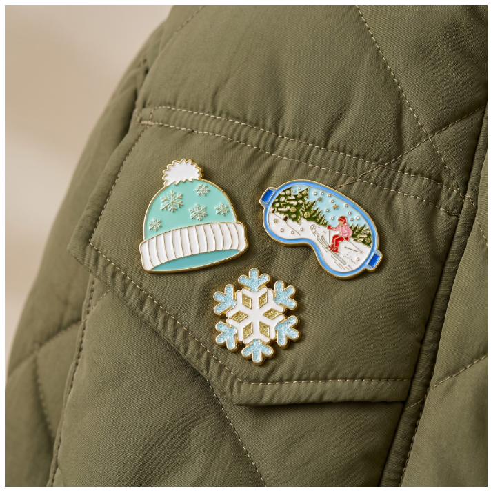 Three winter-themed enamel pins displayed on an olive green quilted jacket: a light blue beanie with white snowflake details, a blue ski mask pin showing a snowy landscape with a skier, and a light blue snowflake pin.