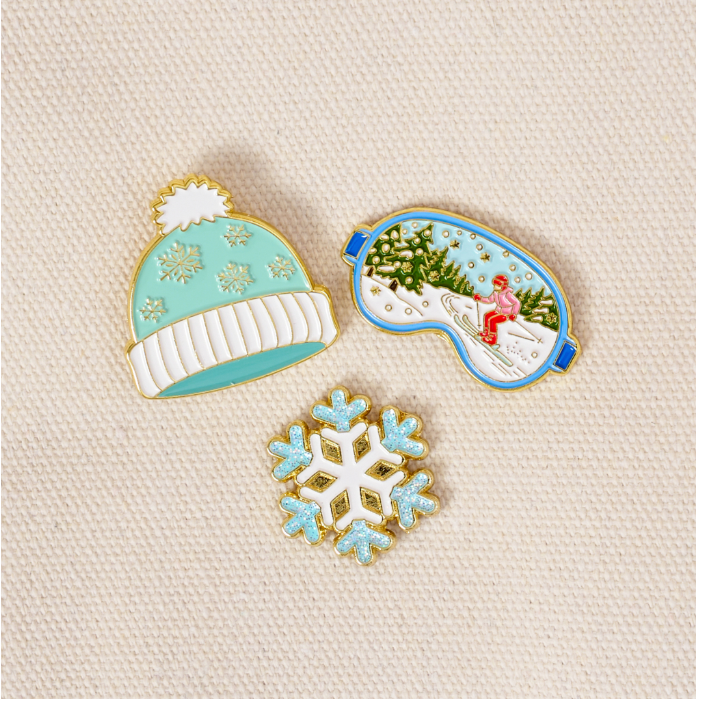 Three enamel pins shaped like a mint green and white winter hat, ski goggles showing a snowy mountain scene, and a snowflake, arranged on a textured cream fabric background.