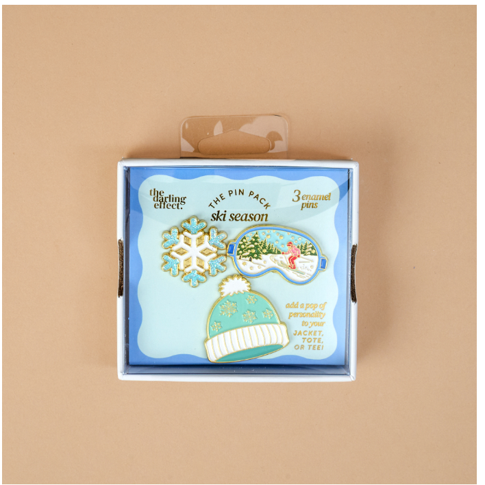 A boxed set of three enamel pins themed around ski season, displayed on a light beige background. The packaging features a light blue background with illustrations of a snowflake, a sleeping landscape pin, and a knit hat pin.
