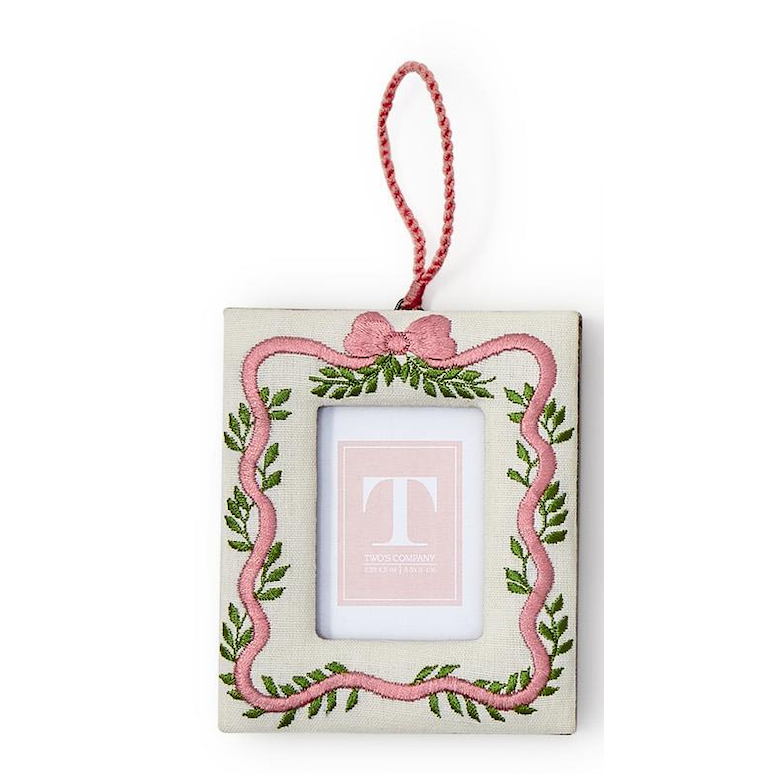 A square fabric ornament with a hanging red cord, featuring a pink embroidered border with green leaf motifs and a pink bow at the top. The center has a white insert with a pink square containing a large letter T.