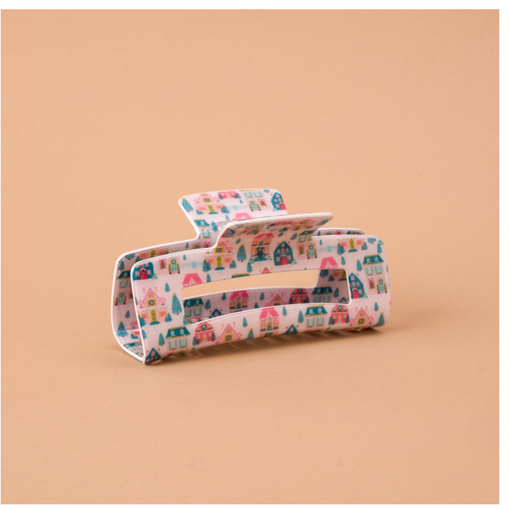A large hair claw clip with a colorful pattern of small houses, trees, and stars on a white background, set against a solid peach-colored backdrop.