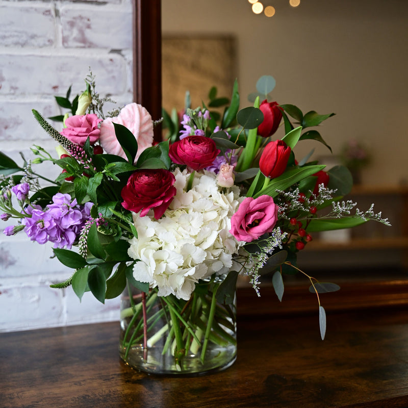 A beautiful floral arrangement featuring hydrangeas, lisianthius, stock, and tulips in a clear round cylinder vase, placed on a wooden surface with a white brick wall in the background.