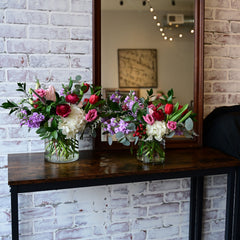 Two floral arrangements to show size difference featuring hydrangeas, lisianthius, stock, and tulips in round cylinder vases on a table against a brick wall backdrop.