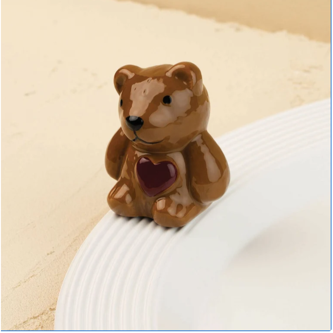 A small glossy brown bear figurine with a dark red heart on its chest, sitting on the edge of a white plate against a warm beige background.