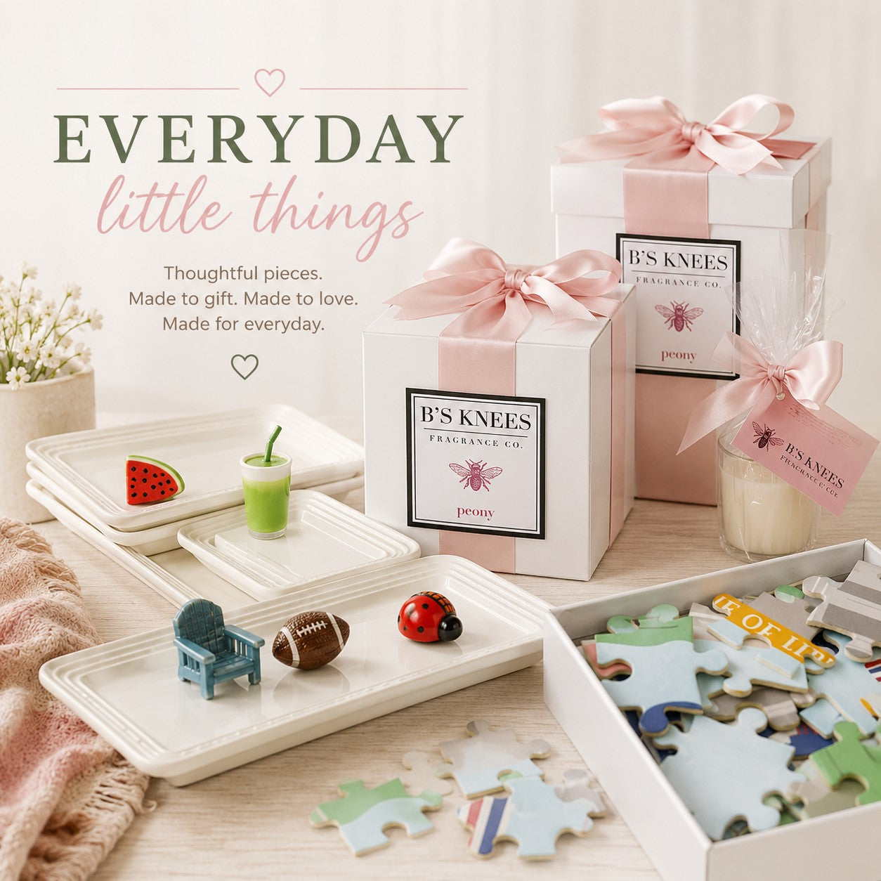 A styled gift set display featuring white gift boxes tied with pale pink ribbons, small decorative items on white trays, and a partially assembled jigsaw puzzle box. Visible branding on the boxes reads 'B'S KNEES' and 'The Knit Co.